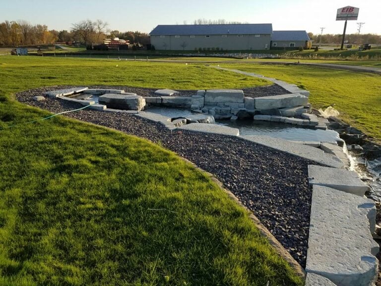 Custom-built decorative water feature with natural stone and gravel pathway in Cedar Rapids, IA. The professionally designed landscape includes a cascading stream surrounded by lush green grass, enhancing the property's curb appeal and drainage management. Expert water feature installation for residential and commercial landscapes.