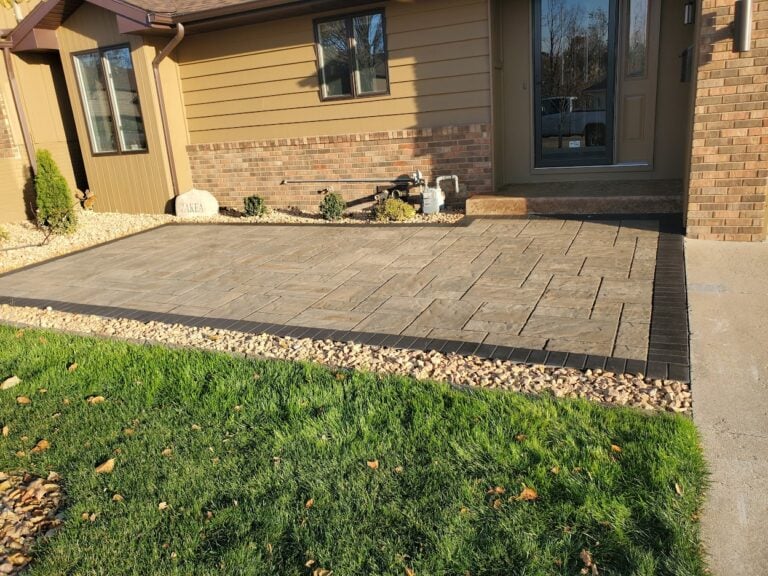 Custom front patio installation in Cedar Rapids, IA, featuring a stamped concrete design with a contrasting border. The professionally landscaped entryway includes decorative rock edging and a well-maintained lawn, enhancing curb appeal and functionality. Expert hardscaping solution for a stylish and durable outdoor space.