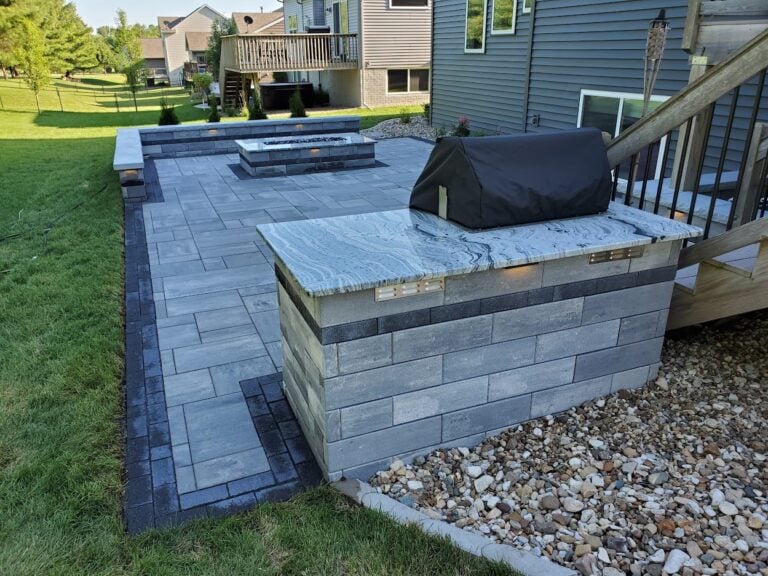 Custom outdoor kitchen with built-in grill and modern paver patio in Cedar Rapids, IA. Featuring a sleek granite countertop, integrated lighting, and a stylish fire pit for the ultimate backyard entertaining space.