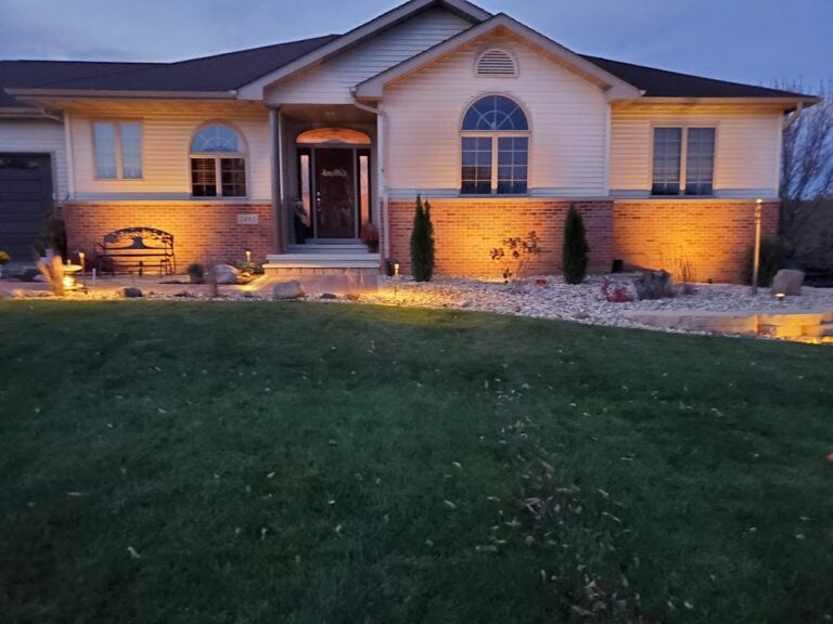 Elegant outdoor landscape lighting installation in Cedar Rapids, IA, highlighting a beautifully maintained front yard and entryway. The professionally designed lighting enhances curb appeal and security, illuminating the home's brick and siding exterior, decorative rock landscaping, and lush green lawn. Expertly crafted for a warm and inviting nighttime ambiance.