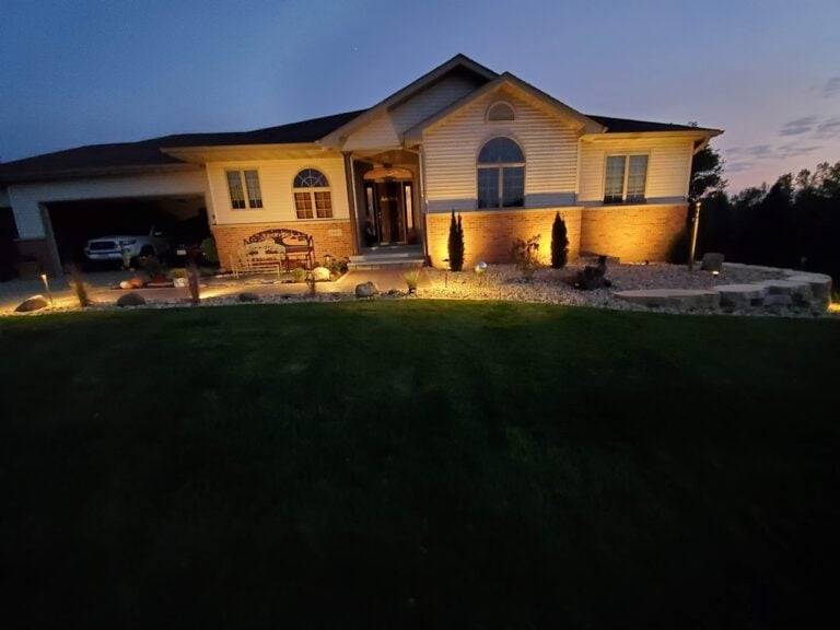 Custom outdoor landscape lighting installation in Cedar Rapids, IA, enhancing a home's front yard with warm accent lighting. The professionally designed illumination highlights the home's brick facade, entryway, decorative rock landscaping, and retaining wall, creating an inviting and elegant nighttime curb appeal.