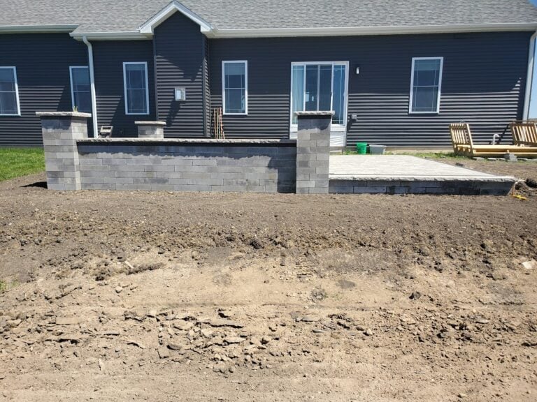 Partially completed outdoor patio with a custom block retaining wall in Cedar Rapids, IA. The hardscape design features a spacious stamped concrete patio and pillar accents, enhancing the backyard of a modern home. Expertly crafted for durability, style, and functional outdoor living space.