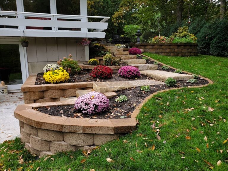 Tiered retaining wall with stone steps and colorful flower beds creating a stunning backyard landscape design. The raised garden beds and hardscaping enhance the outdoor space with beauty and functionality.