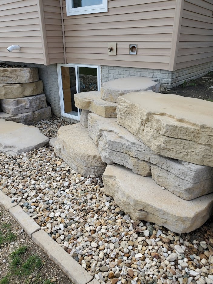 Natural stone staircase with large limestone steps leading to a basement walkout. Custom hardscape design with boulder retaining walls and decorative river rock for erosion control and landscape aesthetics.