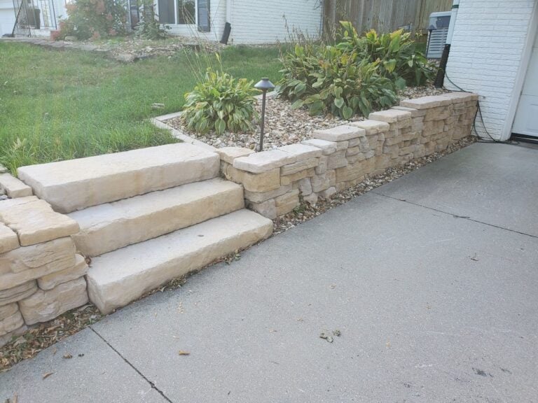 Custom limestone retaining wall and stone step installation in Cedar Rapids, IA, enhancing a home's driveway entrance. The professionally built hardscape features natural stone steps, a decorative rock landscape bed, and integrated pathway lighting for improved accessibility and curb appeal.