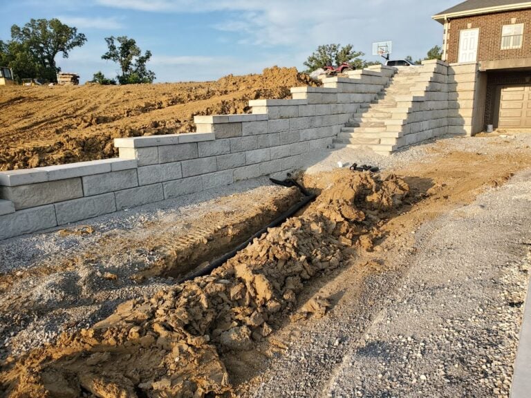 Retaining wall construction in Cedar Rapids, IA, featuring large block wall installation with integrated drainage system. Excavation work in progress for proper water management and erosion control. Professional hardscaping services for residential landscapes.