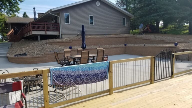 Backyard patio with tiered retaining walls and decorative stone, designed for outdoor entertaining. Features a spacious concrete seating area, modern railing, and elevated deck with access to the home.