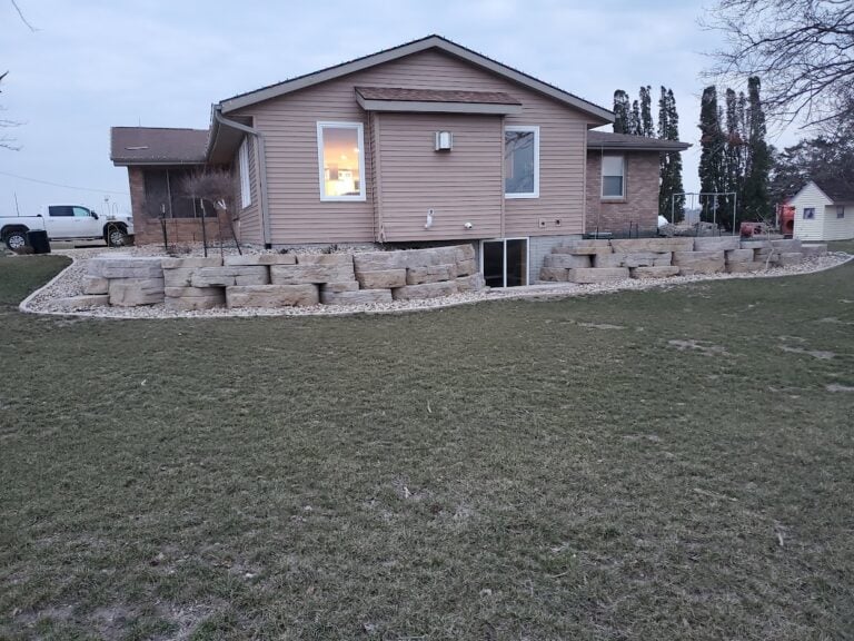 Custom limestone retaining wall installation in Cedar Rapids, IA, designed for erosion control and landscape enhancement. The tiered natural stone wall surrounds a well-maintained home, complemented by decorative rock for low-maintenance landscaping. Expert hardscaping solution to improve curb appeal and property stability.