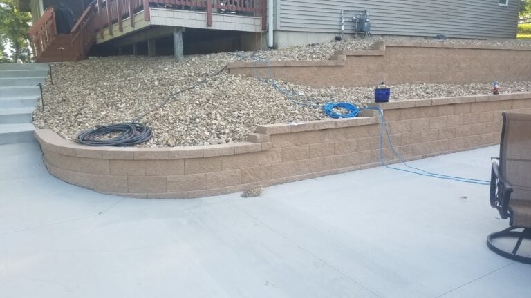 Tiered retaining walls with decorative stone fill, enhancing a backyard patio with erosion control and landscape design. Features a modern concrete patio for outdoor relaxation and entertaining.