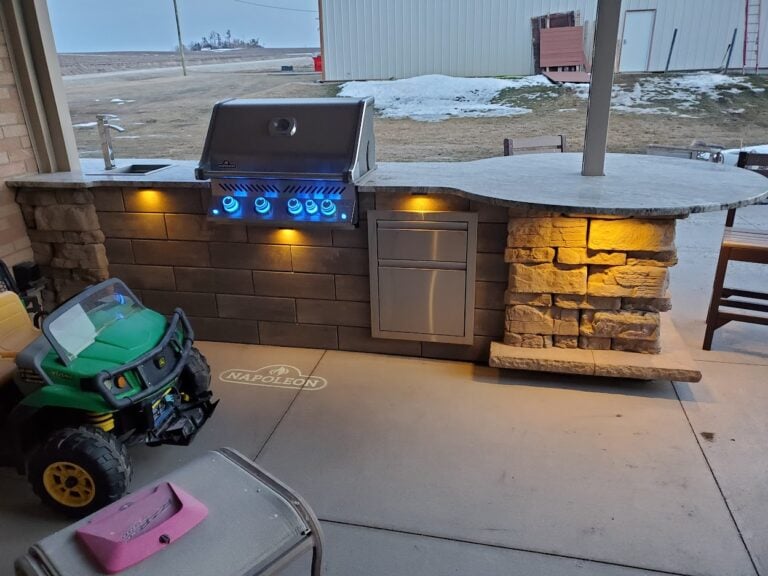 Custom outdoor kitchen with built-in Napoleon grill, stone countertops, and accent lighting in Cedar Rapids, IA. Professionally designed hardscape featuring a stylish grilling station with stacked stone details and integrated storage for an enhanced outdoor cooking experience. Ideal for backyard entertainment and outdoor dining.