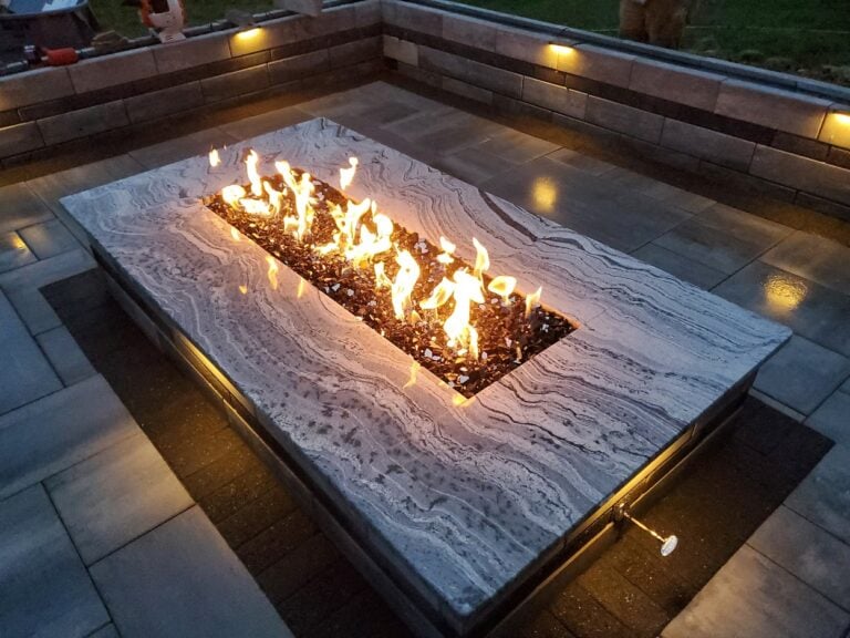 Luxury outdoor fire pit with a sleek stone countertop, built-in lighting, and modern gas burner. This elegant fire feature enhances a custom paver patio, creating a warm and inviting backyard entertainment space.