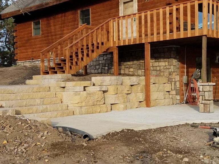 Custom limestone retaining wall and concrete patio installation in Cedar Rapids, IA. The project features large natural stone steps leading to a log cabin-style home with a wooden deck and staircase. Expertly designed hardscape solution for erosion control and enhanced outdoor living space.