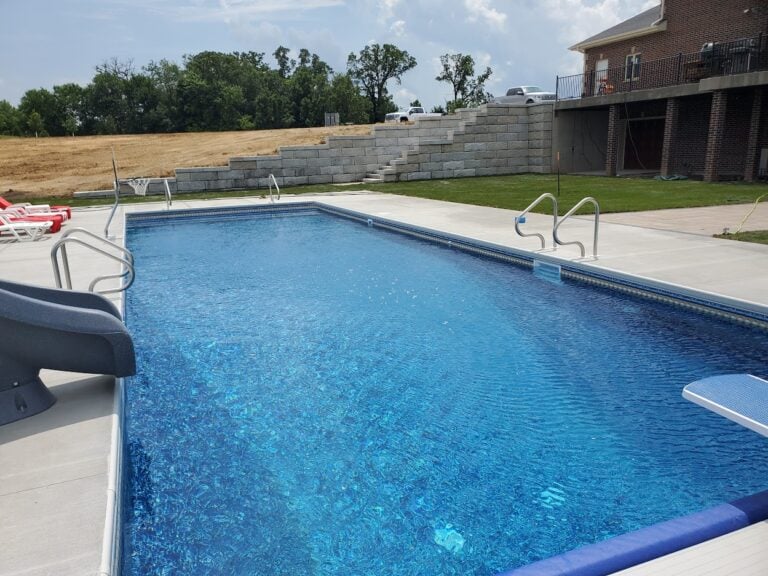 Luxury inground swimming pool with concrete patio and large retaining wall in Cedar Rapids, IA. The professionally designed outdoor space features a custom-built pool, slide, diving board, and tiered block retaining wall for landscape stabilization. Expert poolscape and hardscape installation for a stylish and functional backyard retreat.