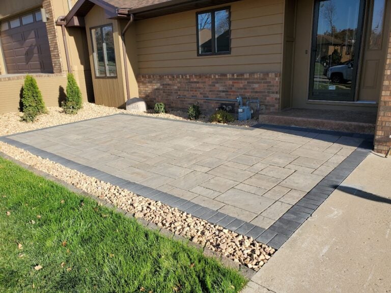 Custom paver front patio installation in Cedar Rapids, IA, featuring a sleek stone design with a contrasting dark border. Surrounded by decorative rock and lush green grass, this professionally crafted hardscape enhances curb appeal and provides a durable, low-maintenance entryway. Expertly designed for style and functionality.