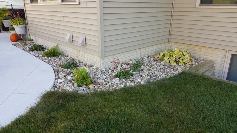 Low-maintenance landscaping with decorative rock and drought-resistant plants around the foundation of a beige siding home. A curved concrete walkway leads to the backyard, bordered by lush green grass. Professionally designed landscape bed for improved drainage and curb appeal in [Target Location, e.g., Cedar Rapids, IA].