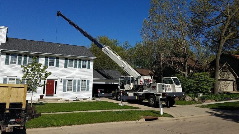Crane lifting materials for a roofing or landscaping project in a residential neighborhood. A large white truck-mounted crane is positioned on a suburban street, assisting with construction work on a two-story home with white siding and black shutters. Blue skies and green trees in the background. Professional heavy equipment service in [Target Location, e.g., Cedar Rapids, IA] for safe and efficient project execution.