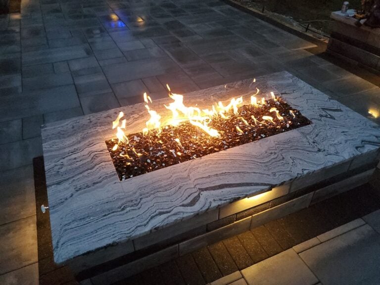 Modern outdoor fire pit with a sleek stone countertop and built-in lighting. This stylish gas fire feature adds warmth and ambiance to a custom paver patio, perfect for backyard entertaining.