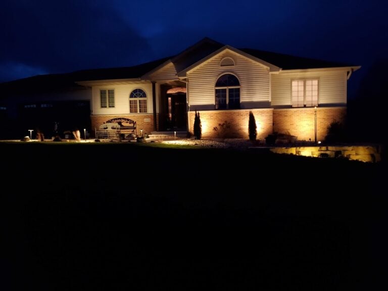 Professional outdoor landscape lighting installation in Cedar Rapids, IA, illuminating a home's exterior, walkway, and decorative rock landscaping. The strategically placed lights enhance curb appeal, improve security, and create a warm, inviting nighttime ambiance. Expertly designed low-voltage lighting for enhanced visibility and elegance.