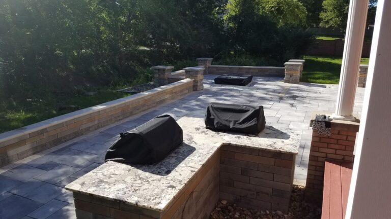 Luxury outdoor kitchen with built-in grills and granite countertops, featuring a spacious paver patio with custom stone seating walls. Perfect for backyard entertaining and outdoor living.