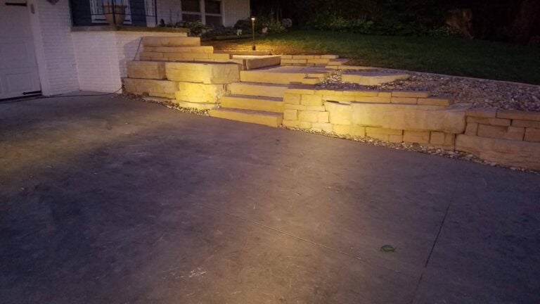 Illuminated natural stone steps and retaining wall leading to a landscaped entryway. Professionally designed hardscape enhances curb appeal, safety, and outdoor aesthetics.
