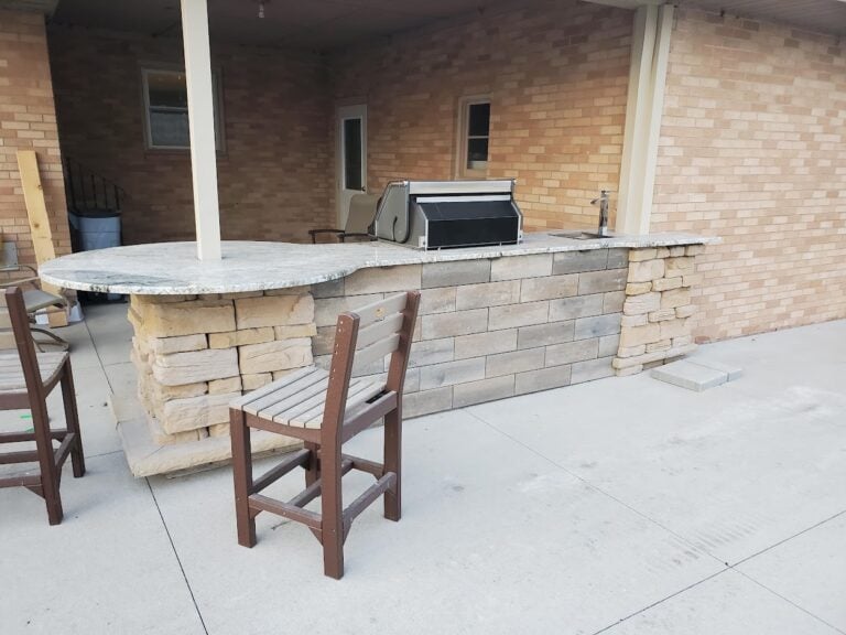 Custom outdoor kitchen with built-in grill, stone veneer base, and granite countertop in Cedar Rapids, IA. The covered patio design includes a spacious bar-height seating area, perfect for outdoor dining and entertaining. Professionally installed hardscape solution for a stylish and functional backyard space.