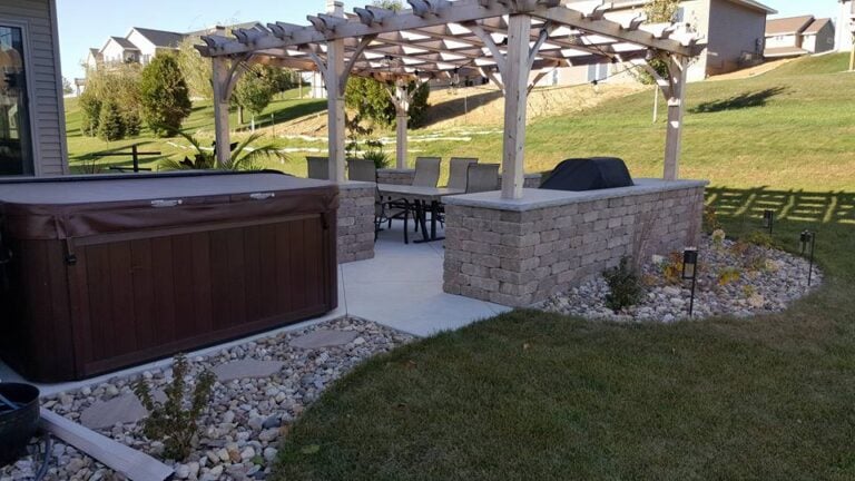 "Custom outdoor living space in Cedar Rapids, IA, featuring a pergola-covered patio with a paver seating wall, a built-in grill area, and a relaxing hot tub. The professionally designed landscape includes decorative rock, pathway lighting, and lush green grass, creating the perfect backyard retreat for entertaining and relaxation."