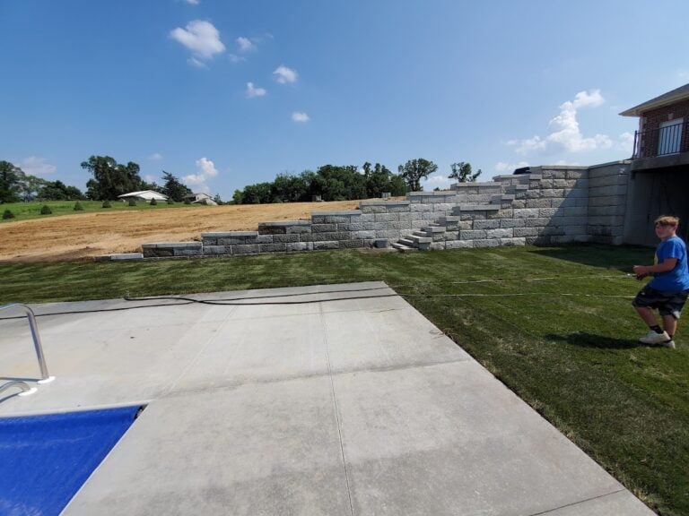 Custom retaining wall installation in Cedar Rapids, IA, featuring tiered concrete blocks for slope stabilization and landscape enhancement. Fresh sod and a newly poured concrete patio create a functional outdoor space beside a swimming pool. Expert hardscaping and grading services for residential backyards.