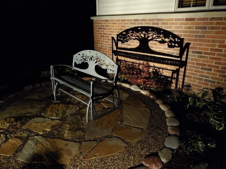 Beautifully illuminated flagstone patio with decorative stone edging in Cedar Rapids, IA. The custom outdoor seating area features a metal bench with a tree design, casting a striking shadow against the brick exterior. Professionally designed landscape lighting enhances the ambiance and highlights the intricate details of this cozy garden retreat.