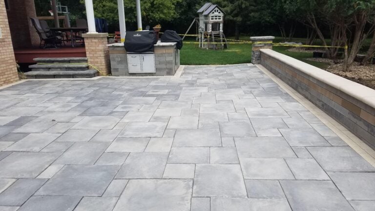 Modern backyard patio with gray paver flooring, built-in outdoor kitchen, and seating wall. Designed for entertaining, this spacious outdoor living space features a sleek hardscape layout with easy access to the deck and lawn.