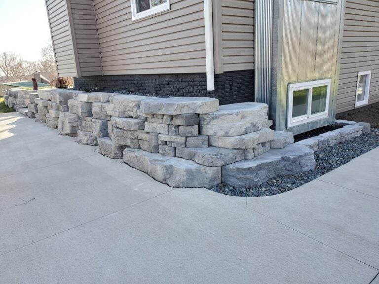 Tiered natural stone retaining wall with integrated steps, complementing a modern home exterior. Designed for structural support and aesthetic appeal in a well-maintained landscape.