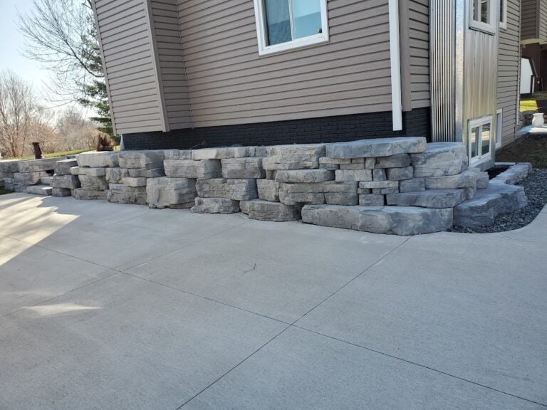 Natural stone retaining wall alongside a concrete driveway, enhancing curb appeal and erosion control. Expertly crafted for durability and seamless landscape integration.