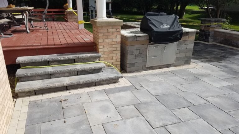 Backyard patio with built-in outdoor kitchen and stone steps leading to a redwood deck. Featuring modern gray pavers, integrated grill station, and hardscape design for entertaining. Perfect for outdoor cooking and relaxation.